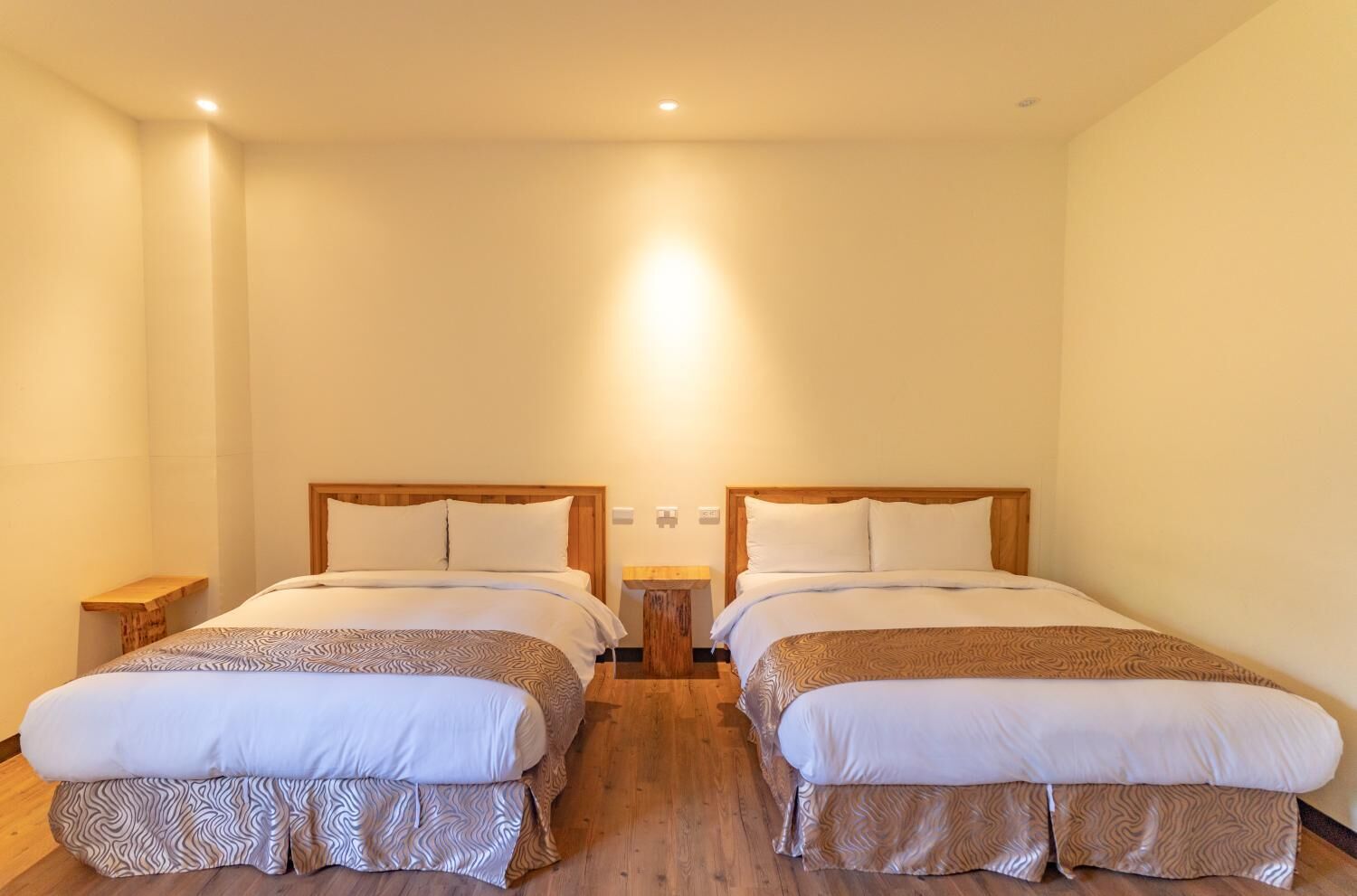 Camera d'albergo con due letti, testiere in legno, biancheria bianca e un piccolo comodino con una lampada.