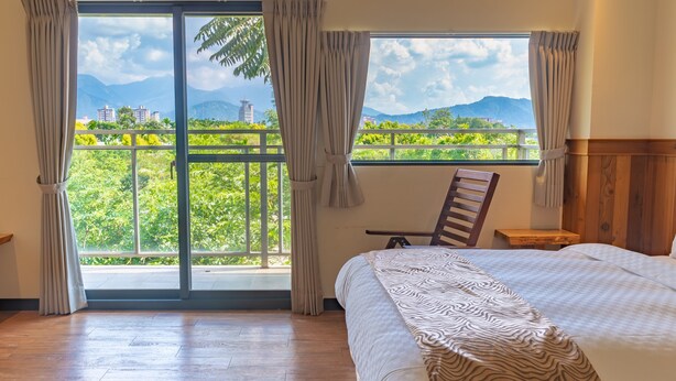 A bedroom with a bed, a chair, and a view of greenery and buildings through a window. - Lane 523 (Puli)