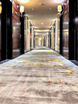 Un pasillo de hotel con alfombra estampada, puertas a ambos lados y lámparas fijadas a la pared.