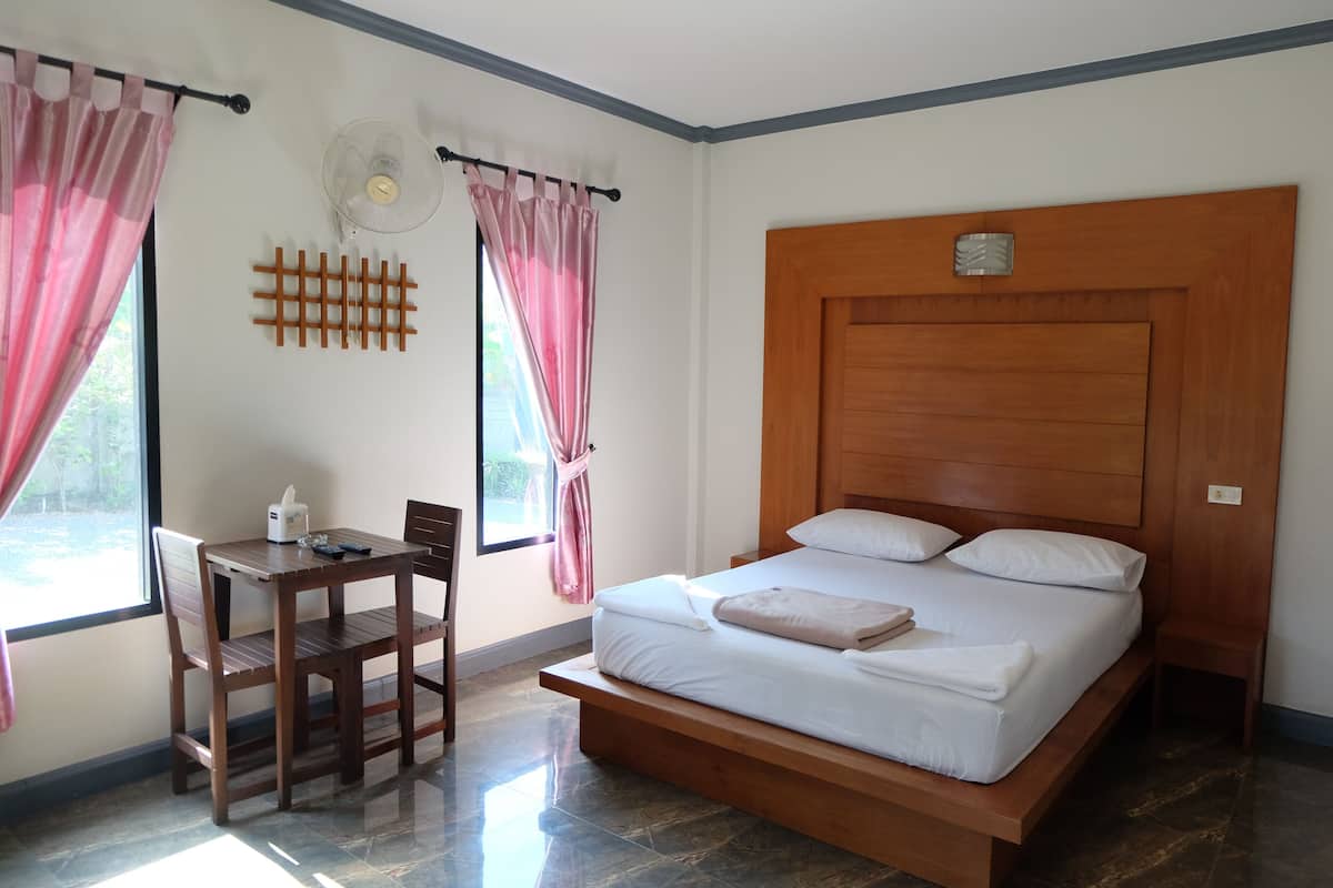 Un dormitorio con cabecera de madera, cama con ropa blanca, mesita de noche, una silla y un escritorio con un ventilador y una tetera.