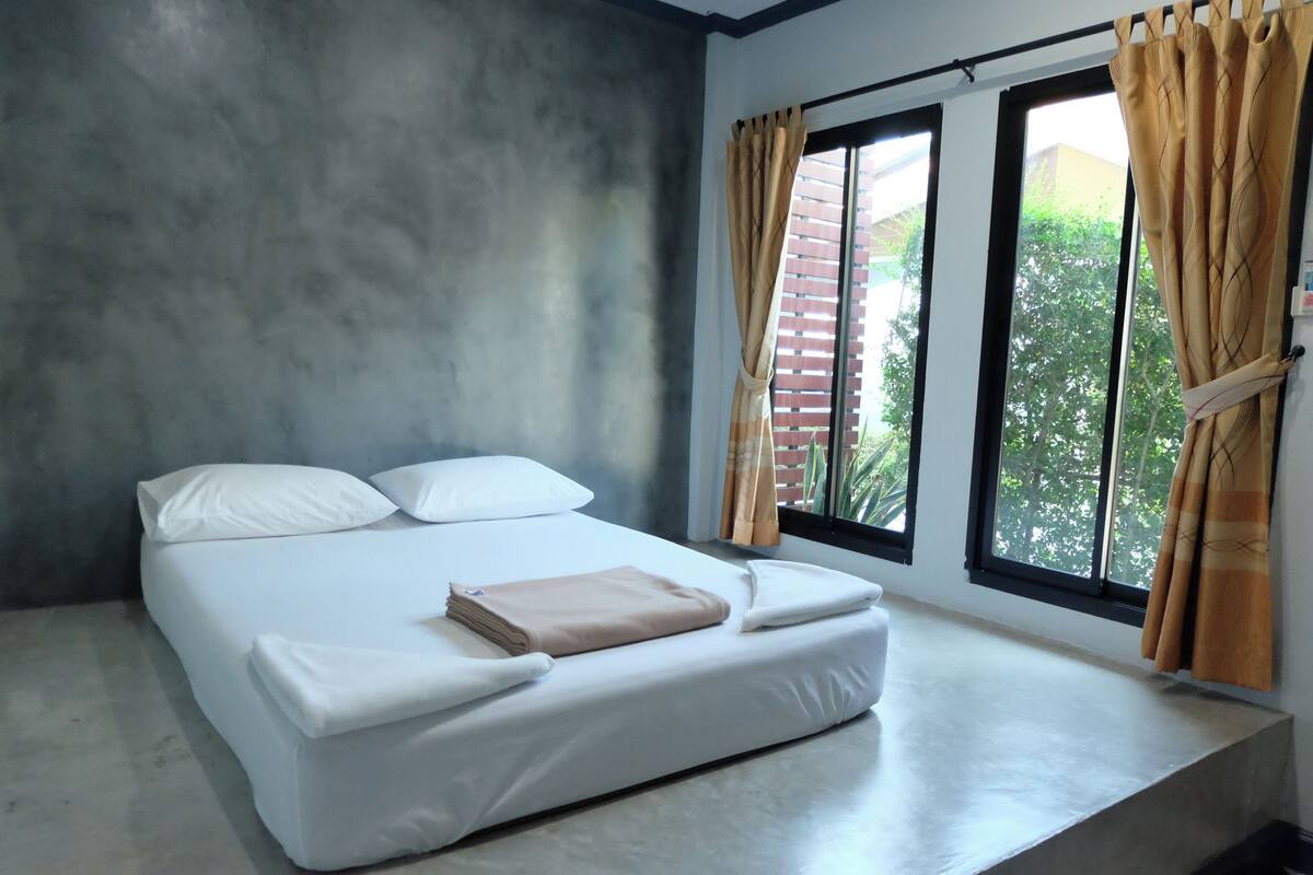 Una cama individual con ropa de cama blanca, una colcha color beige y dos almohadas. Hay una ventana con cortinas y vistas al exterior con vegetación.