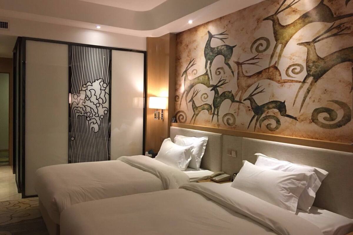 Une chambre d’hôtel avec un grand lit, deux oreillers, une table de chevet avec une lampe et une fresque murale représentant des cerfs.
