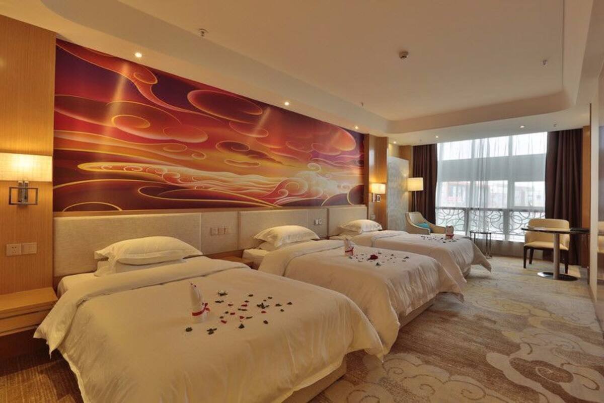 Une chambre d’hôtel avec trois lits, une grande fresque abstraite sur le mur, une vue sur la ville à travers de grandes fenêtres et une petite table avec des chaises.