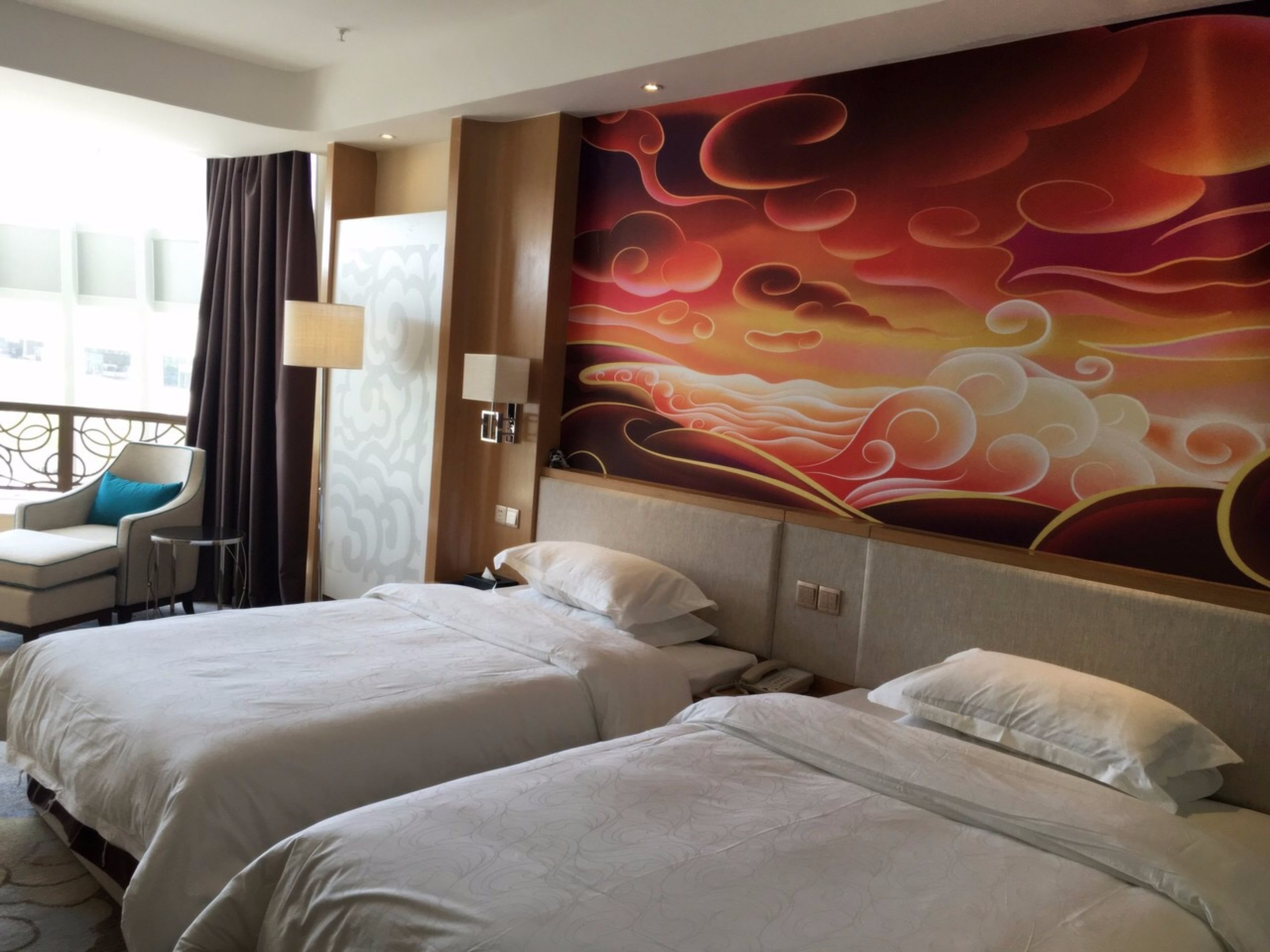 Een hotelkamer met twee bedden, een groot muurschildering, een zithoek met een stoel en een tafel, en een raam met gordijnen.