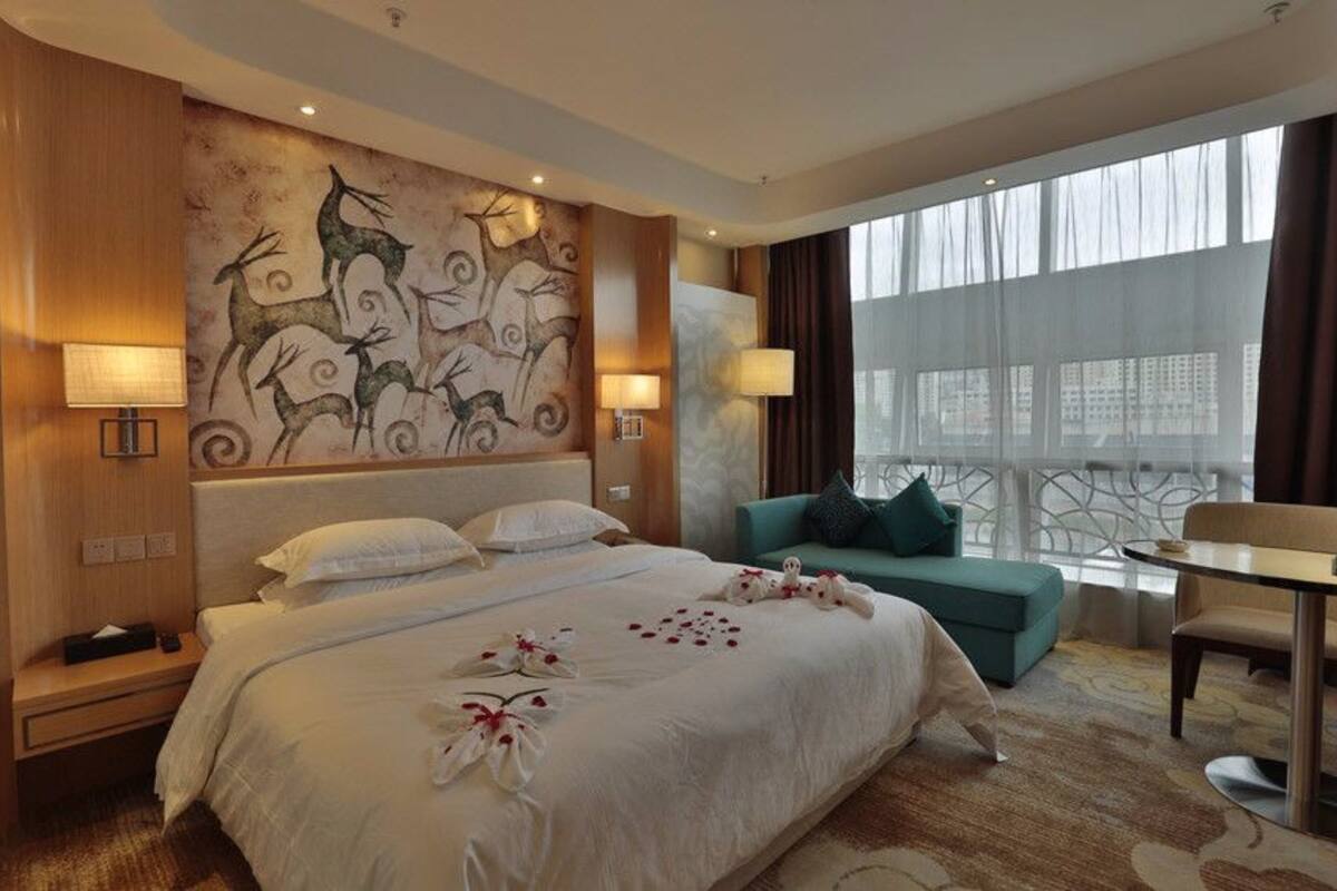 Une chambre d’hôtel avec un grand lit, un canapé, une table à manger et une vue sur les bâtiments, visible à travers de grandes fenêtres.
