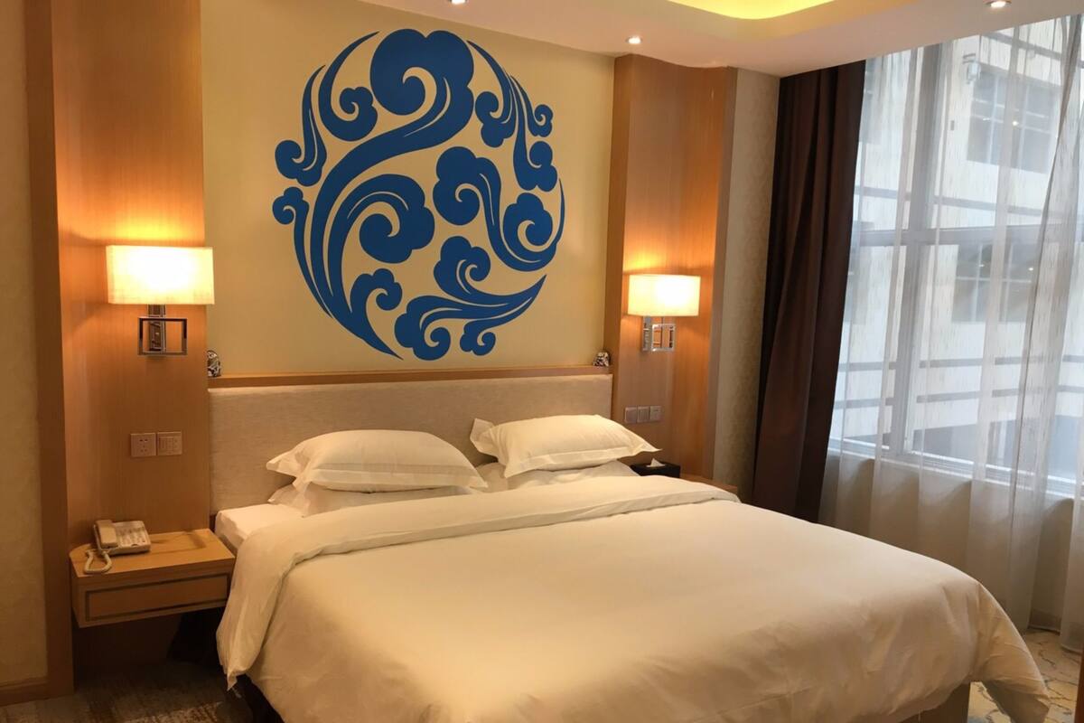 Une chambre d’hôtel avec un grand lit, deux lampes de chevet, un élément décoratif mural et une fenêtre avec des rideaux.