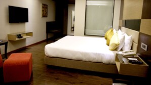 A hotel room with a large bed, a TV mounted on the wall, a desk with a telephone, and a window with blinds. - Hotel Shakti Continental (Angul)