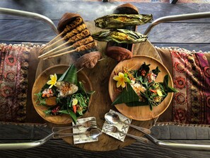 A wooden table with a variety of dishes, including skewers, rice, and vegetables, set on a patterned tablecloth. - Taman Nauli Bed & Breakfast (Tibubeneng)