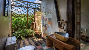 A bathroom with a rustic design, featuring a wooden sink, a shower with a colorful tile wall, and a view of lush greenery outside. - Taman Nauli Bed & Breakfast (Tibubeneng)