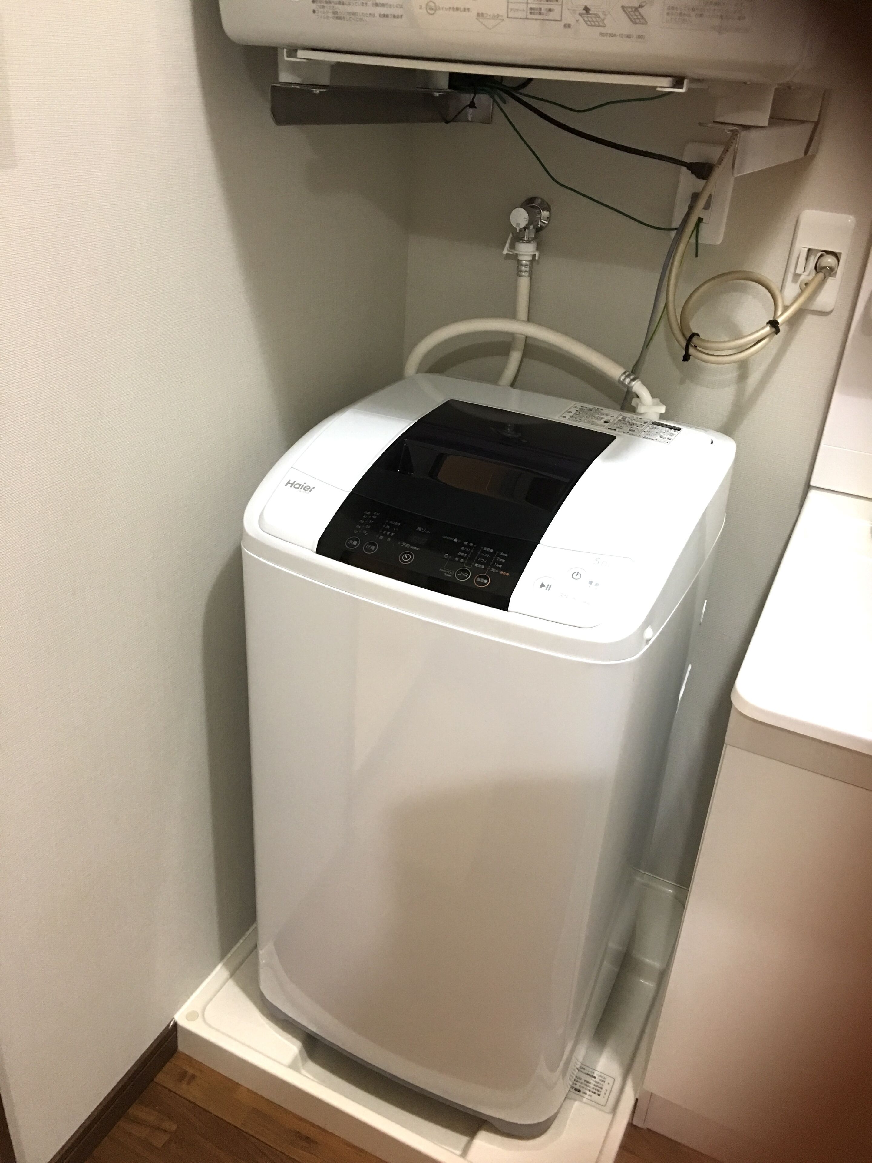 A compact, white, wall-mounted water heater with a control panel, located in a corner of a room with wooden flooring and a white wall.