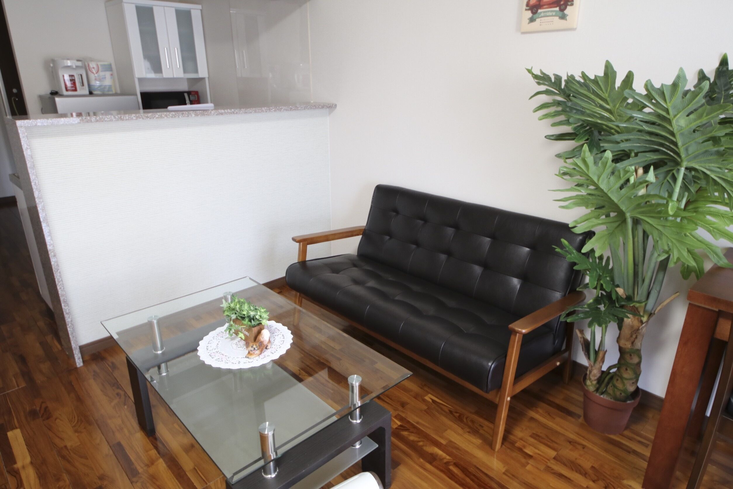 Un salón con una mesa de centro de vidrio, un sofá de cuero negro, una planta en maceta y una zona de cocina al fondo.