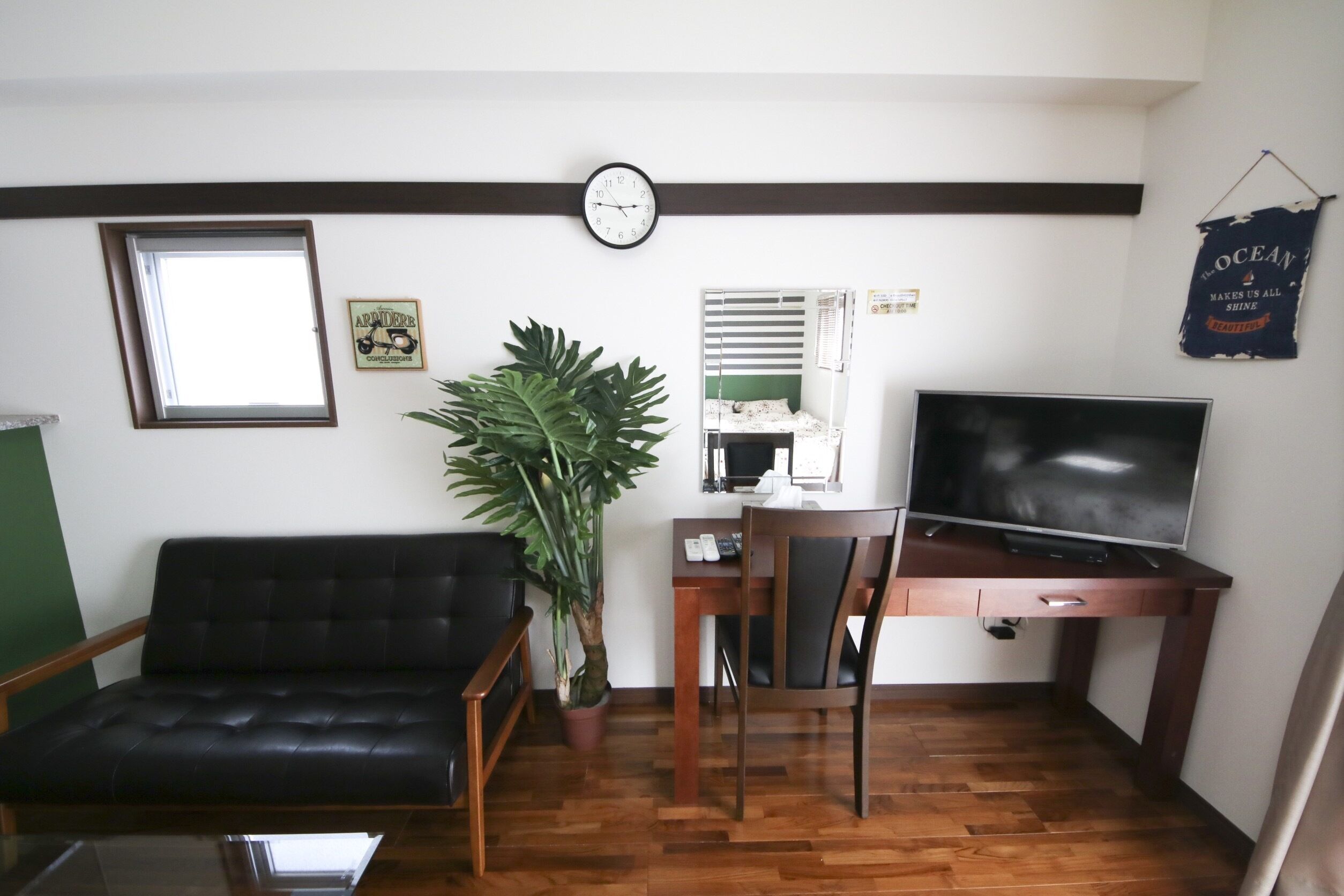 Una sala de estar con un sofá de cuero negro, un escritorio de madera con un televisor, una planta en maceta y un reloj en la pared.