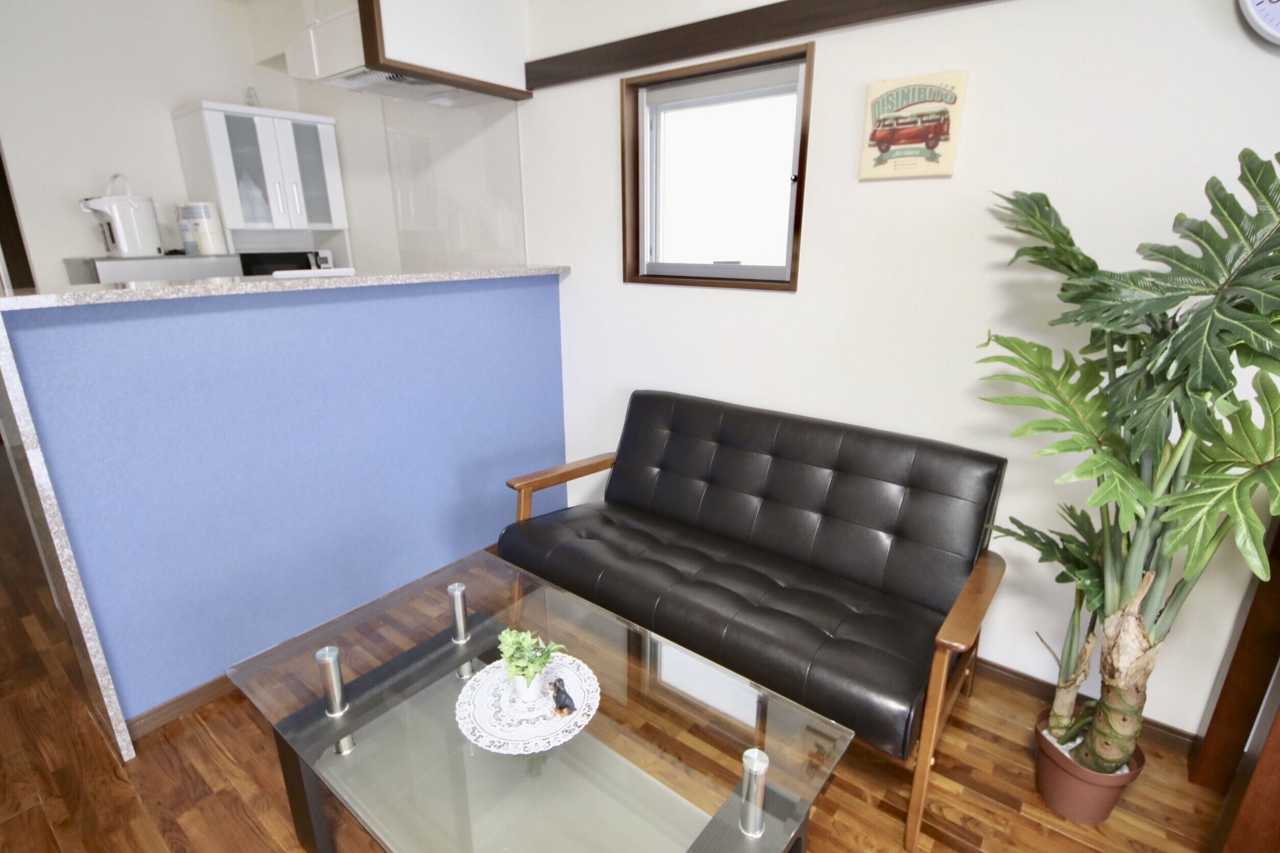 Una sala de estar con un sofá de cuero negro, una mesa de centro de vidrio con un plato decorativo, una planta en maceta y una zona de cocina con armarios y un microondas.
