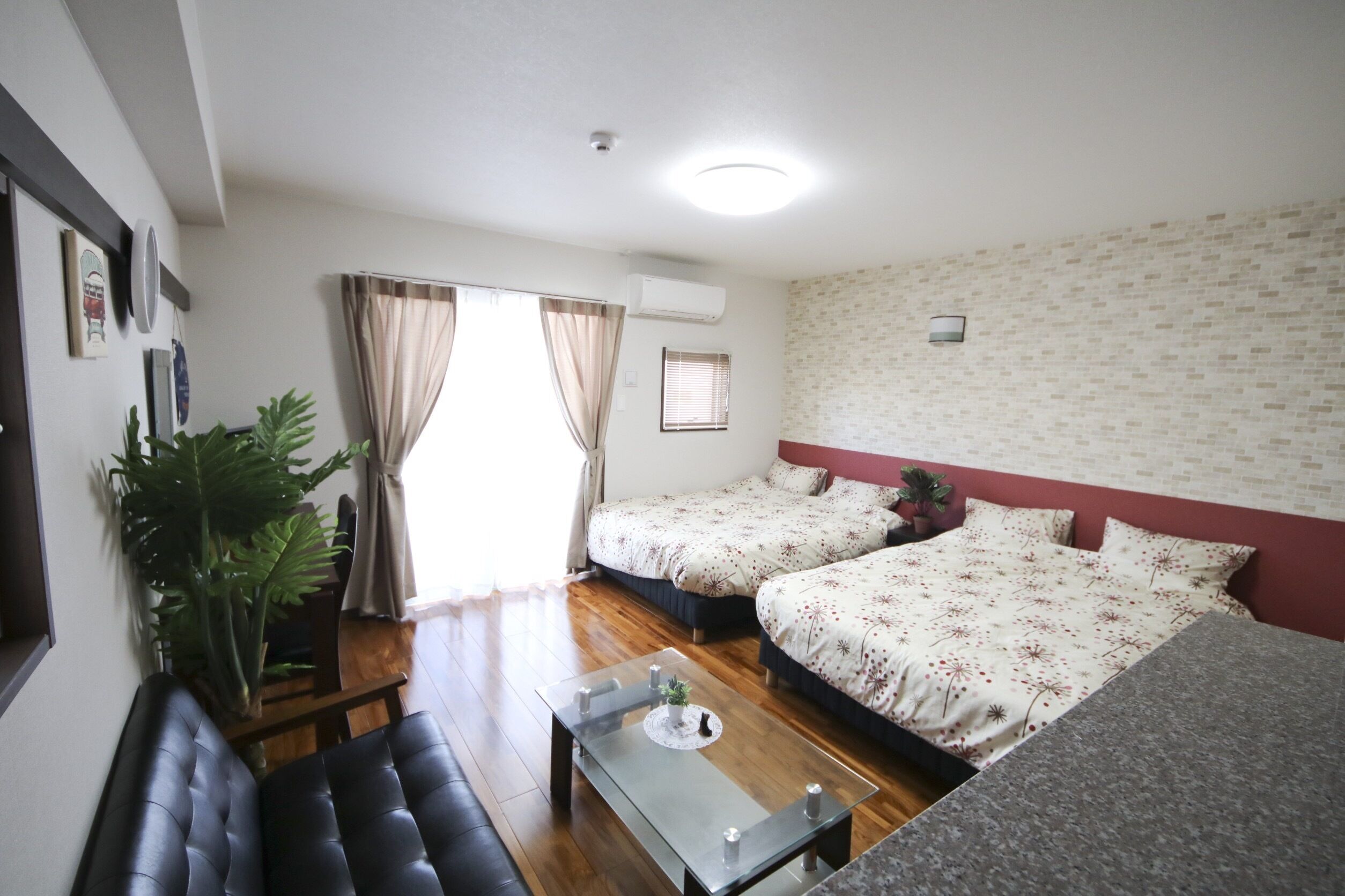 A room with two beds, a dining table, a large potted plant, and a window with curtains.