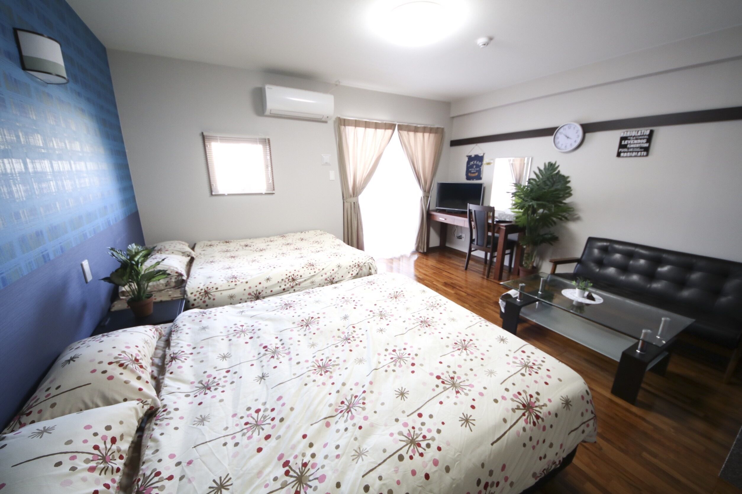 Habitació d'hotel amb dos llits, una taula de treball, un sofà, una televisió i un rellotge a la paret.