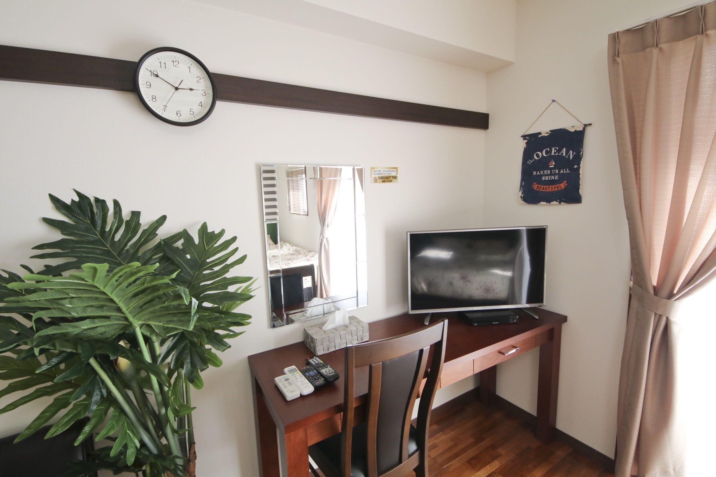 A room with a wooden desk, a television, a clock, a mirror, and a large plant.