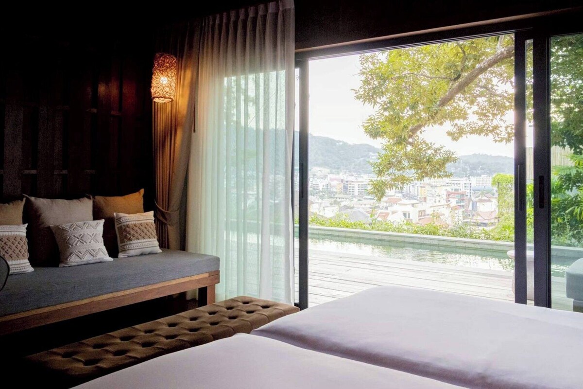 A bedroom with a bed, a sofa, and a view of a cityscape through large windows.