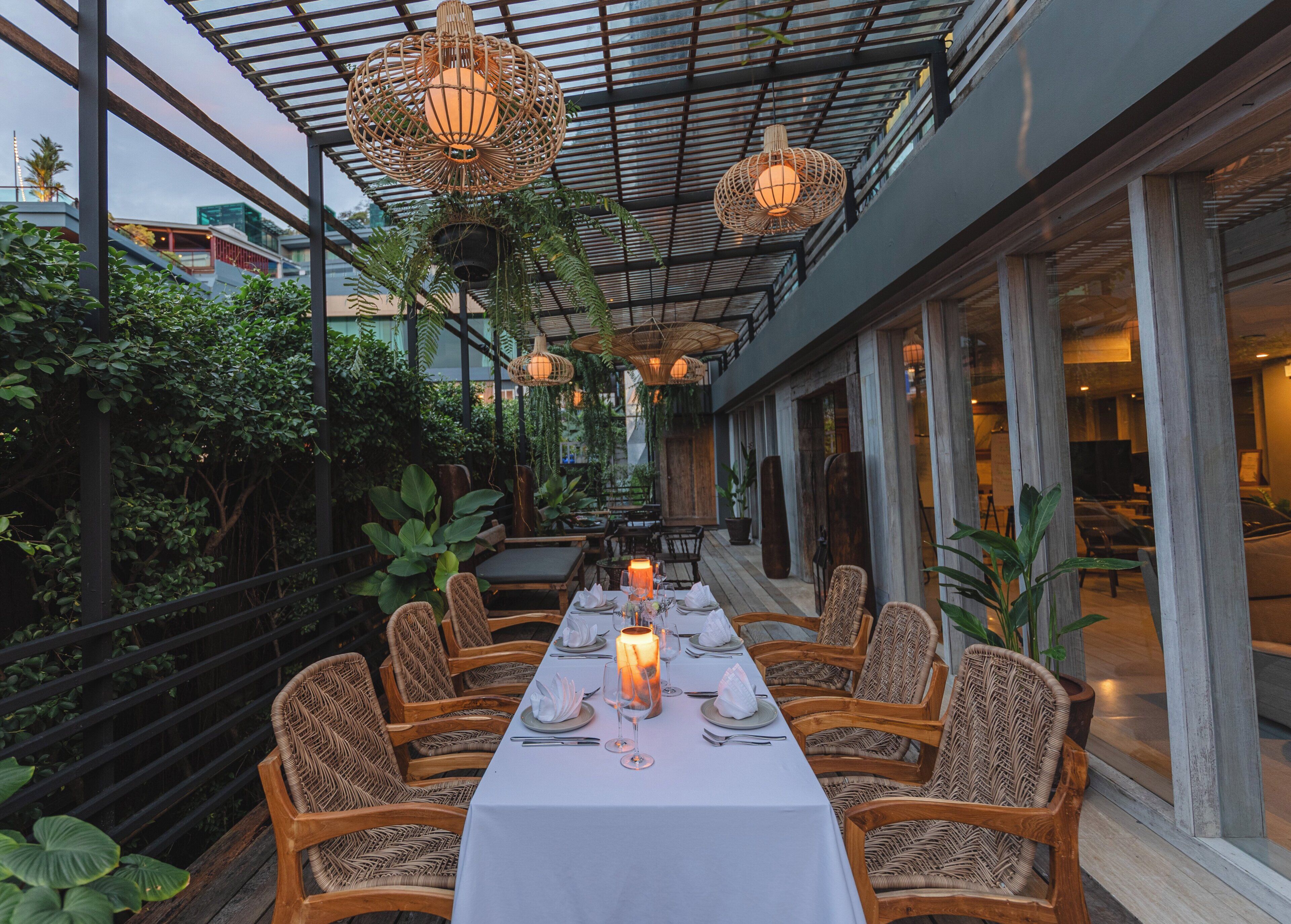 An outdoor dining area with wicker chairs and a long table set for a meal, surrounded by greenery and under a covered structure.