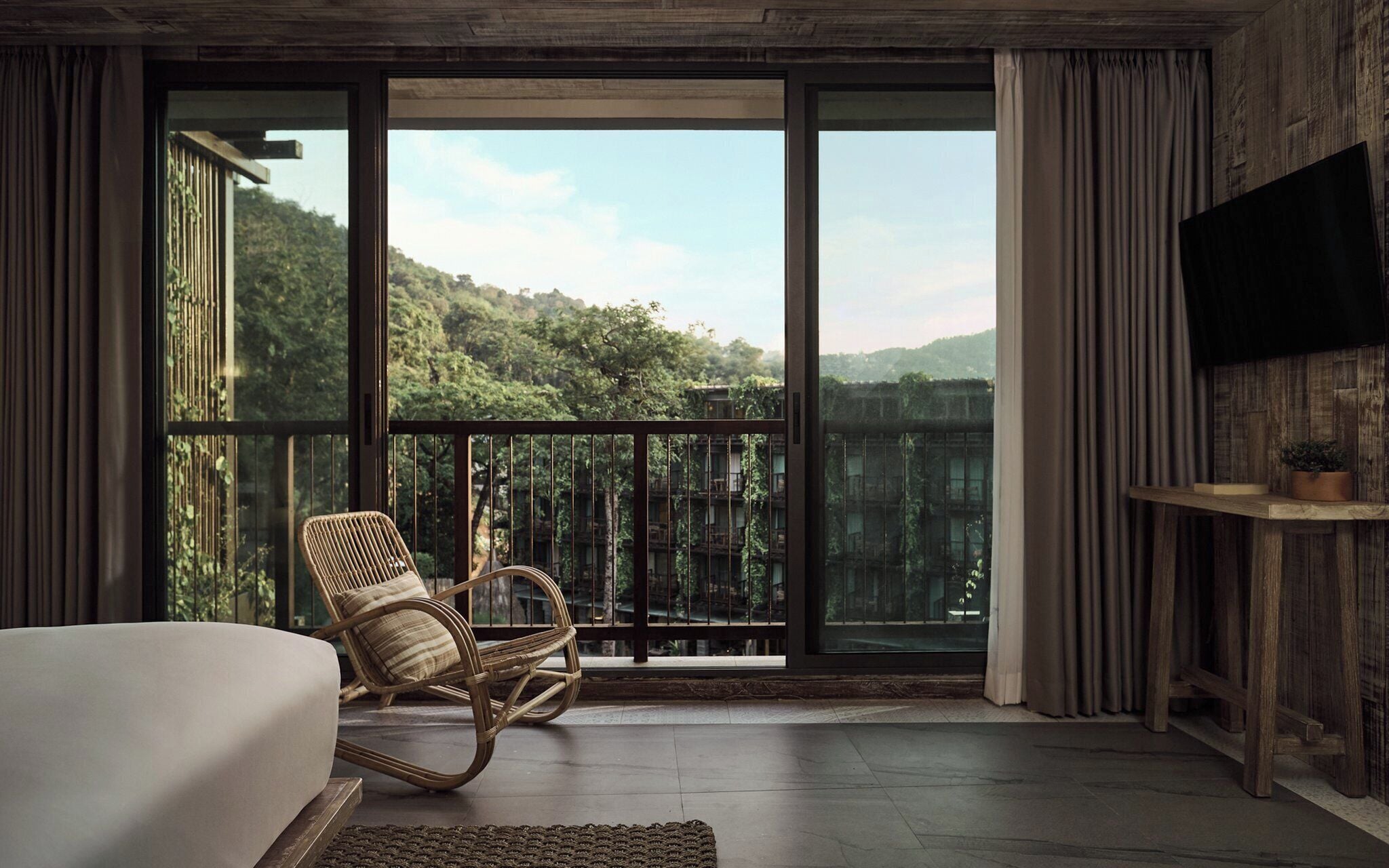 A bedroom with a large window, a bed, a wicker chair, and a TV on the wall.