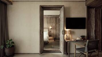 A hotel room with a wooden door leading to a bathroom, a bed, a desk with a lamp, and a television on the wall.