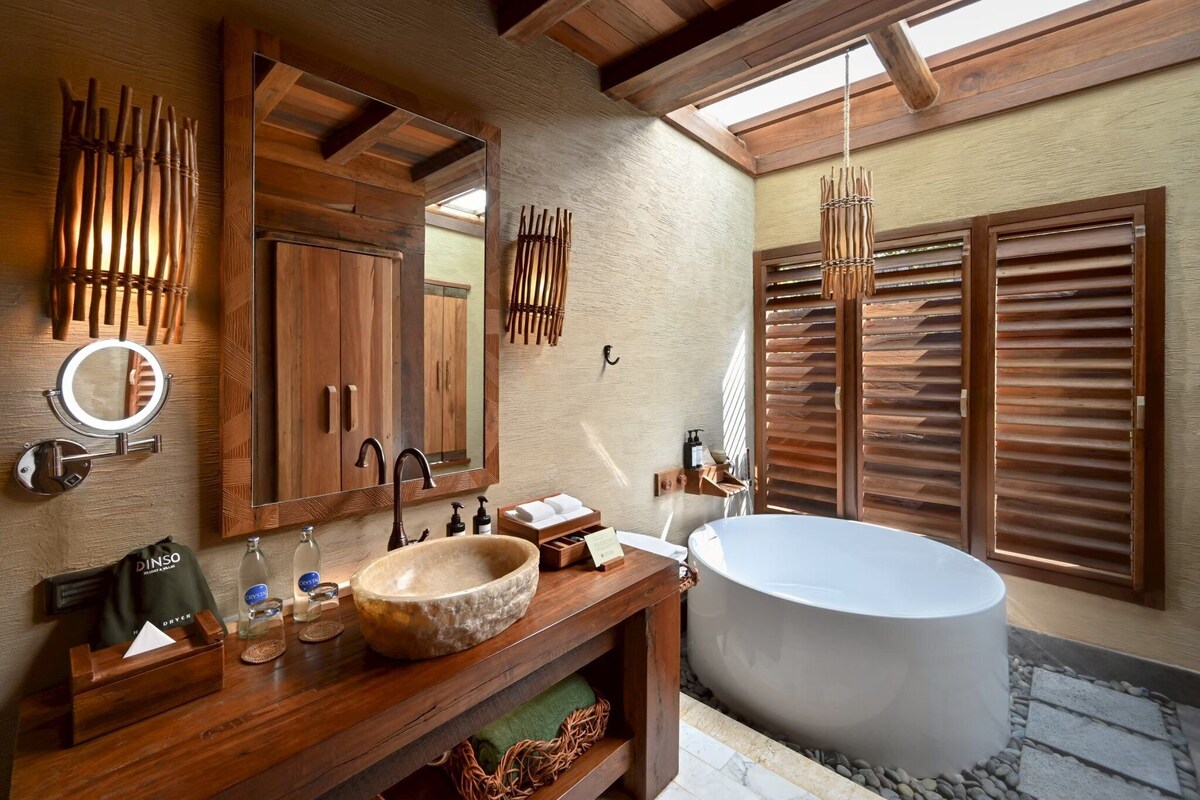 A bathroom with a wooden vanity, a large round sink, a bathtub, wooden slats, and a hanging light fixture.