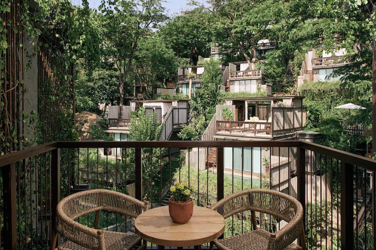 A balcony with wicker chairs and a table, surrounded by lush greenery and wooden structures.