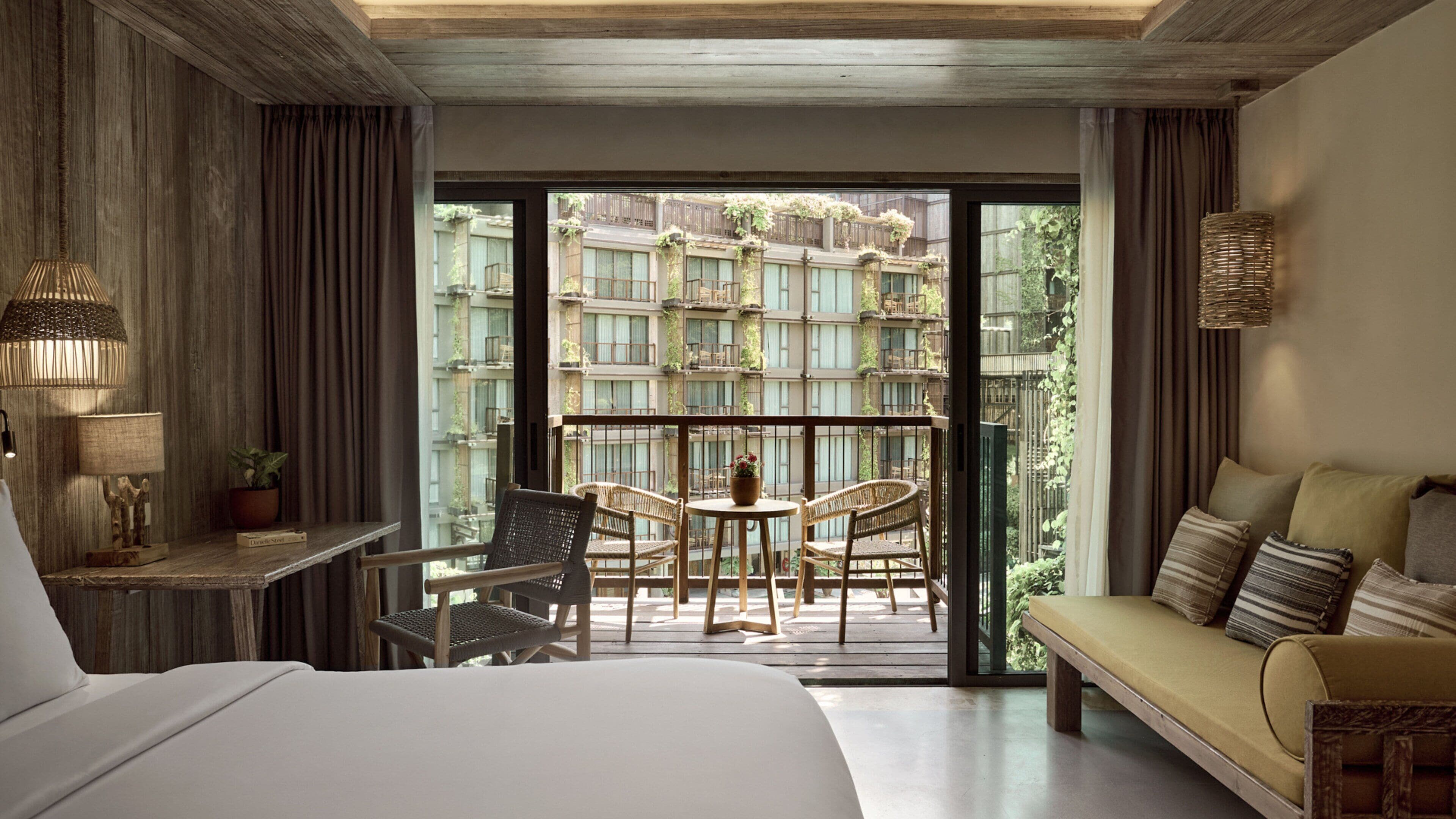 A hotel room with a bed, a sofa, a table with chairs, a lamp, and a view of a courtyard with plants and buildings.