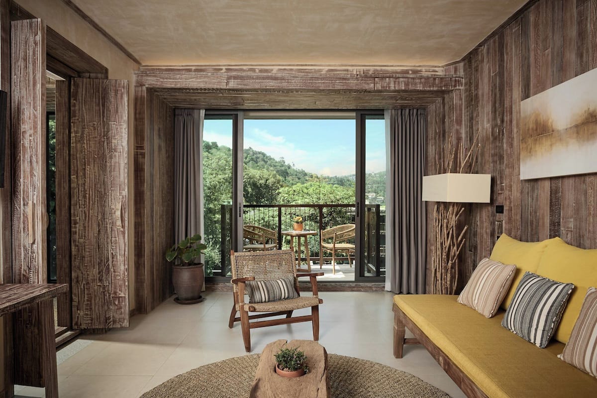 A spacious room with a large sliding glass door leading to a balcony with a view of greenery. There is a wooden chair, a sofa with striped cushions, and a small table with a plant.