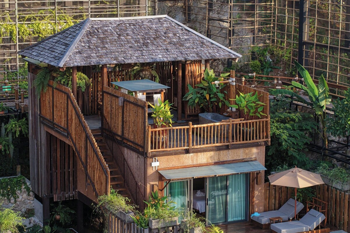 Une maison en bois à deux étages, dotée d’un balcon, d’une piscine et d’une végétation luxuriante.