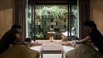 Une salle de spa équipée de deux tables de massage, d’une petite table en bois ornée d’une statuette d’oiseau, et offrant une vue sur la verdure grâce à un mur de verre.