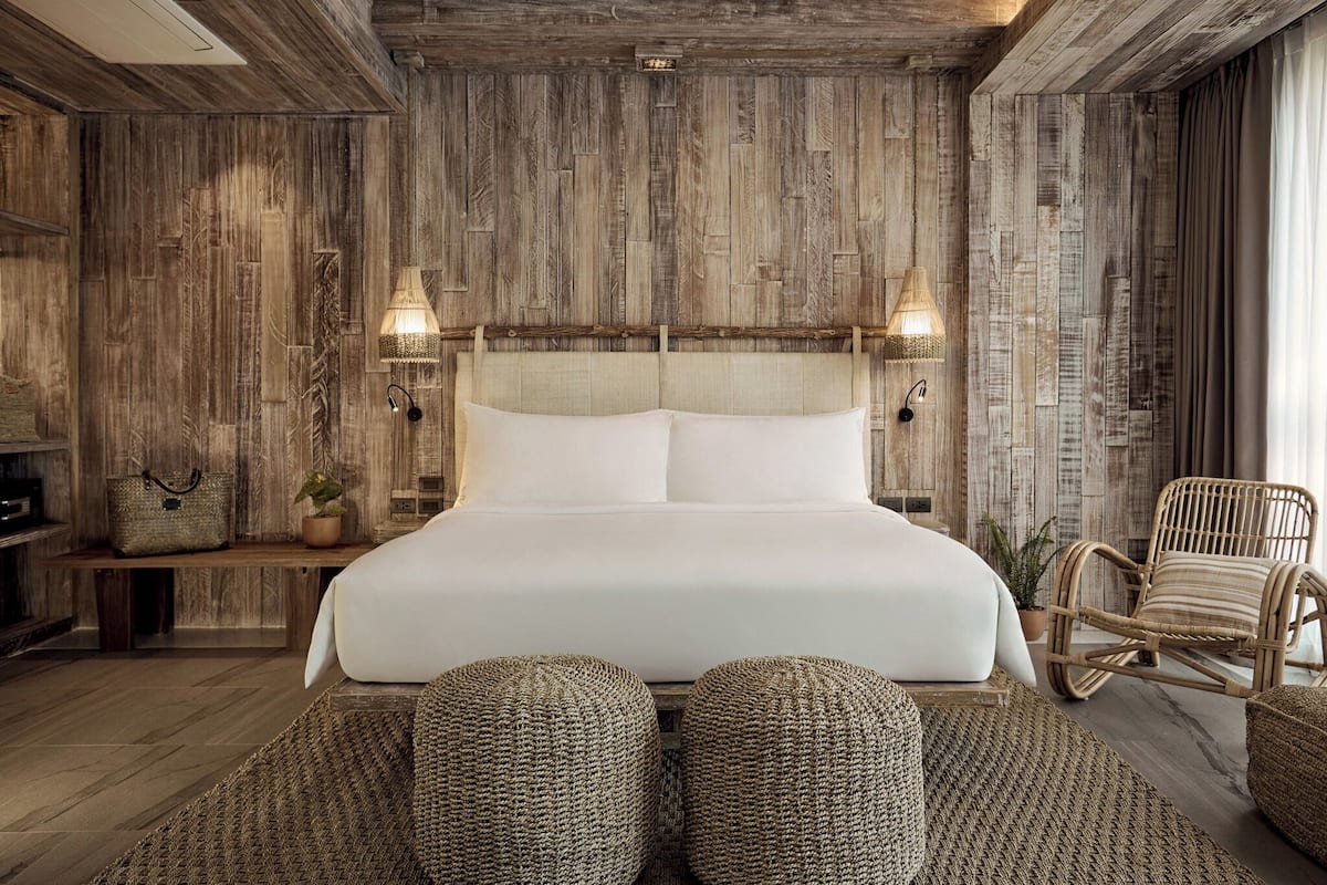 A bedroom with a wooden wall panel, a bed with white bedding, two wall-mounted lamps, a wicker chair, and a bench with a handbag and plant.
