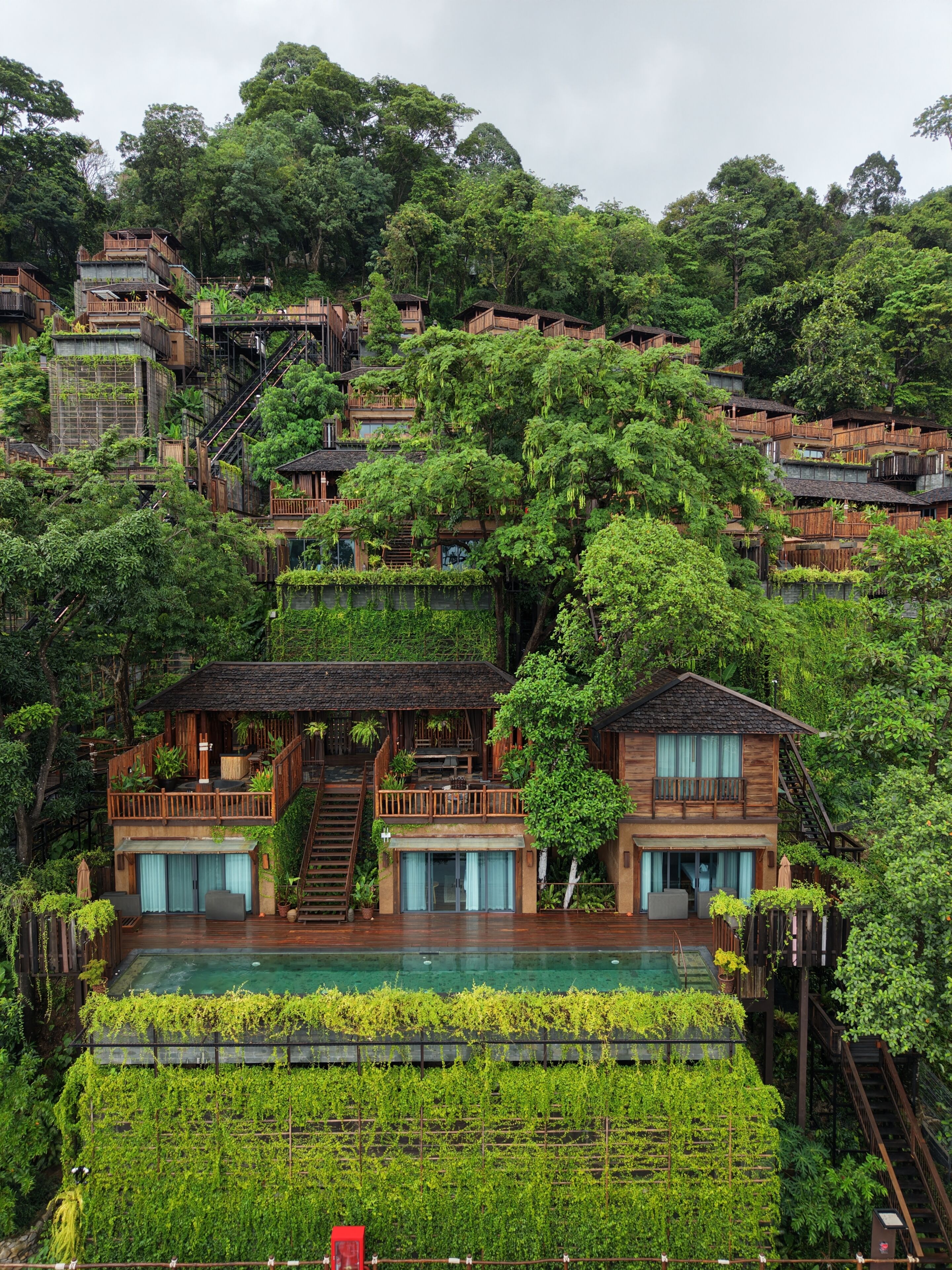 A multi-story wooden house with a balcony, surrounded by lush greenery and a swimming pool.