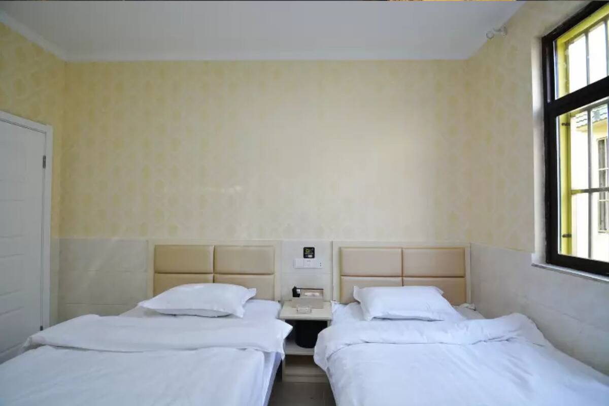 A hotel room with two single beds, a small bedside table with a lamp, and a window with a view of the outside.
