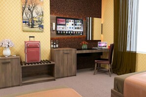 Ein Hotelzimmer mit Schreibtisch, Laptop, Stuhl, Bett, an der Wand montiertem Fernseher und einem kleinen Tisch mit Blumenvase.