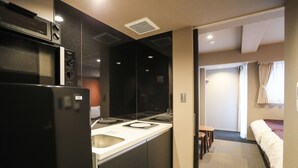 A compact kitchen with a refrigerator, microwave, and stove. A small dining area with a table and chairs is visible. The room has a neutral color scheme and a view of a bedroom with a bed and curtains.
