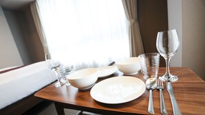 A neatly set dining table with white plates, bowls, cutlery, and glasses, positioned near a window with curtains.