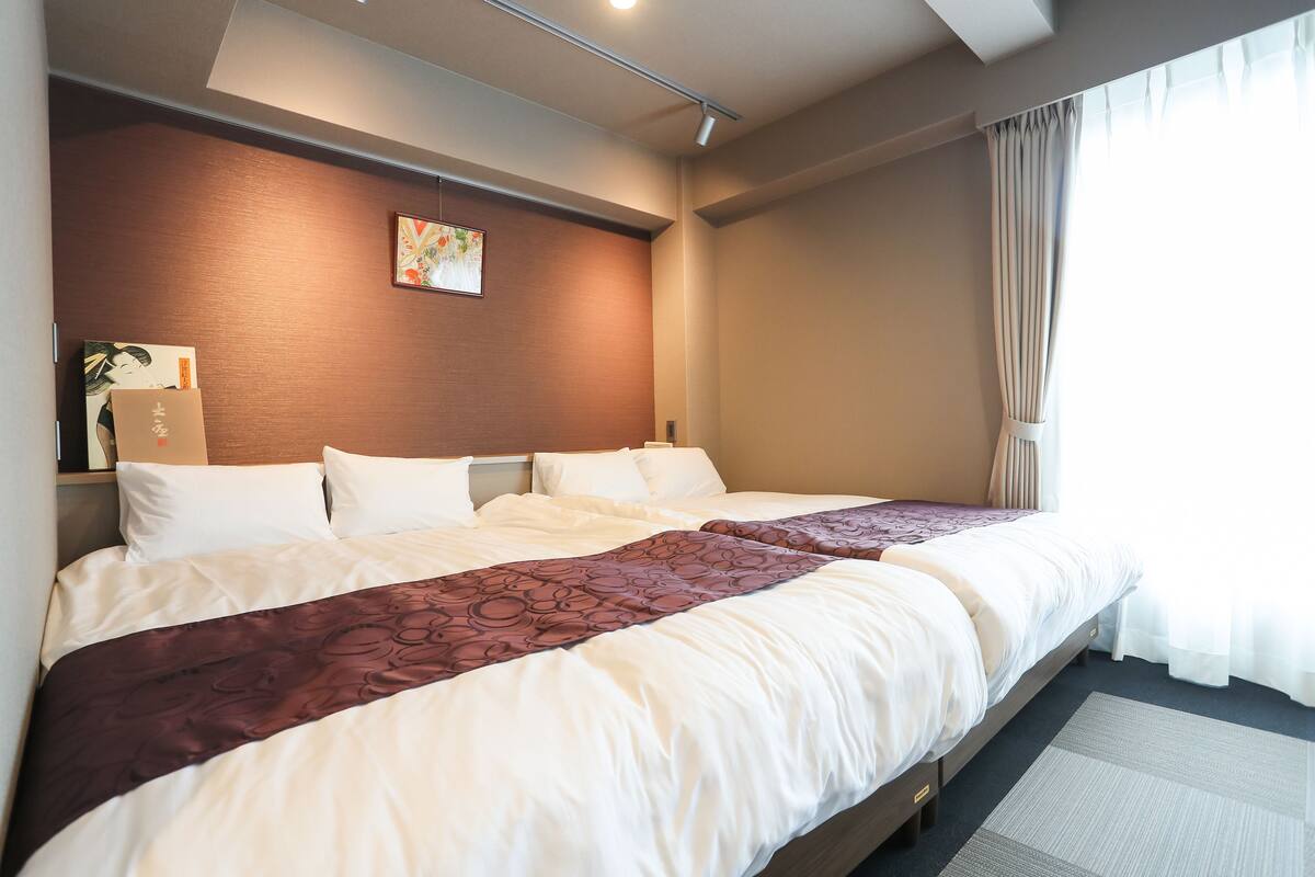 Una habitación de hotel con una cama grande, cabecero, mesita de noche con lámpara y ventana con cortinas.