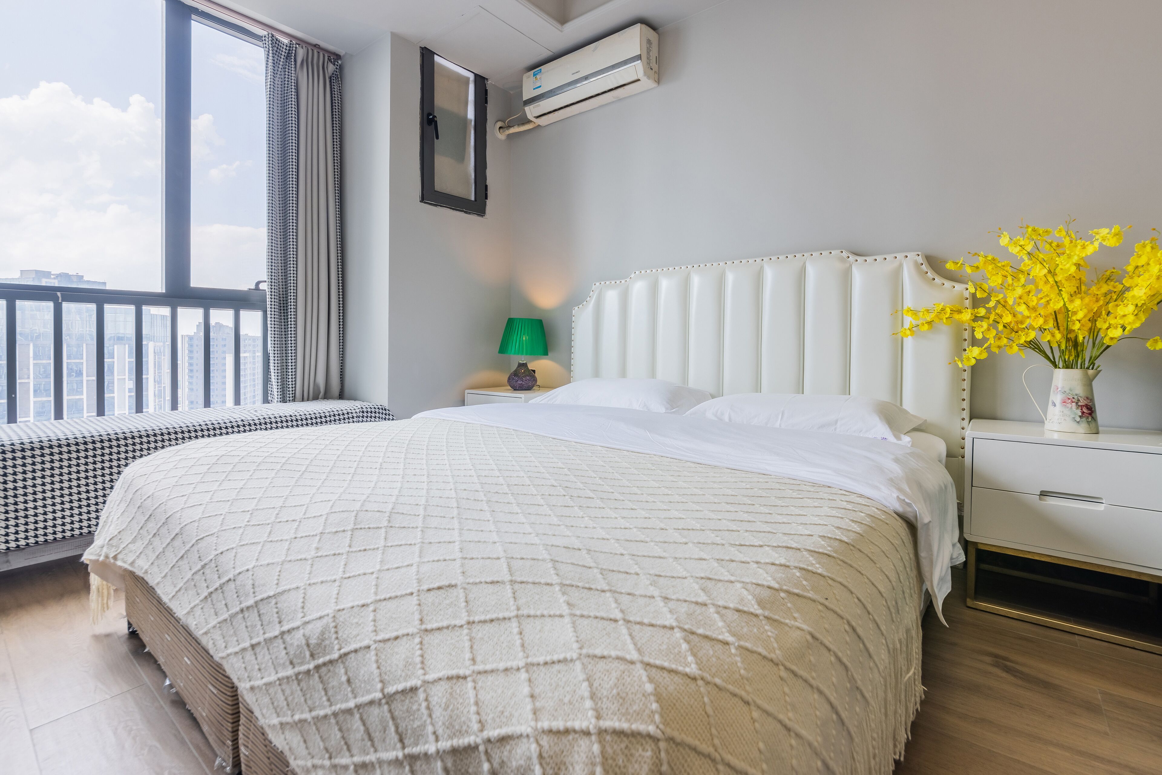 A hotel room with a large bed, a bedside table with a lamp, a vase with flowers, and a view of the city through the window.
