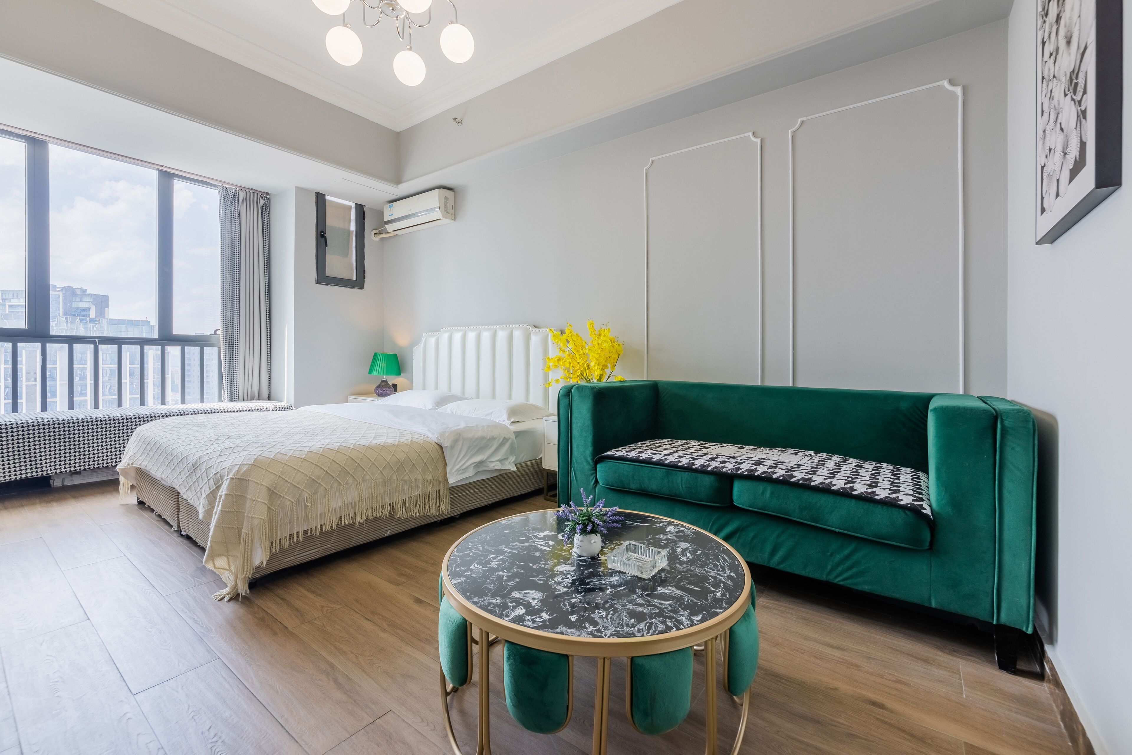 A modern room with a bed, a green sofa, a round table, and a view through large windows.