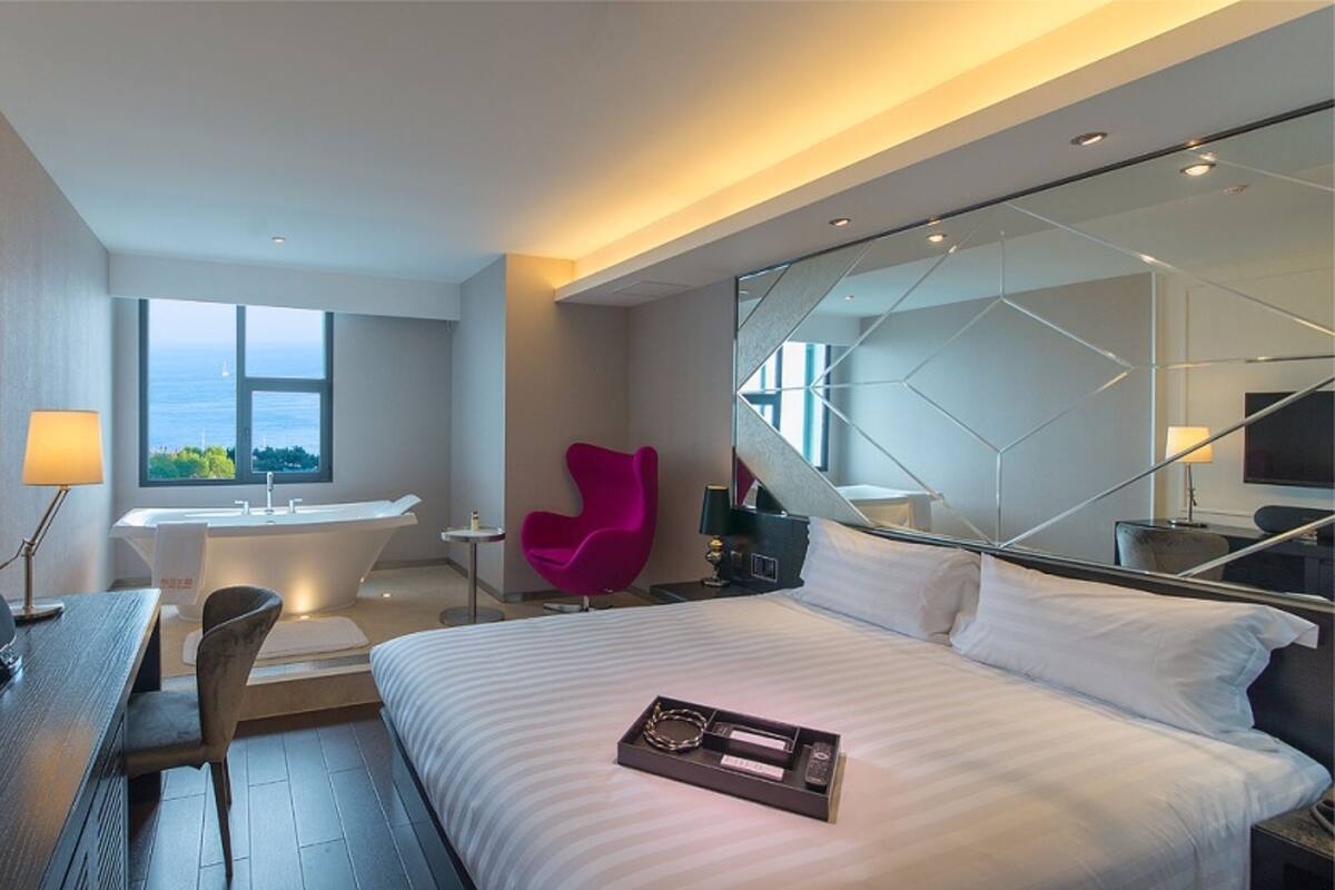 Una habitación de hotel moderna con una cama grande, un escritorio con una silla, una mesita, una silla rosa, una bañera y un ventanal con vista al mar.