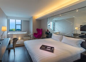 A modern hotel room with a large bed, a desk with a chair, a small table, a pink chair, a bathtub, and a large window with a view of the sea. - Crystal Orange Hotel (Qingdao)