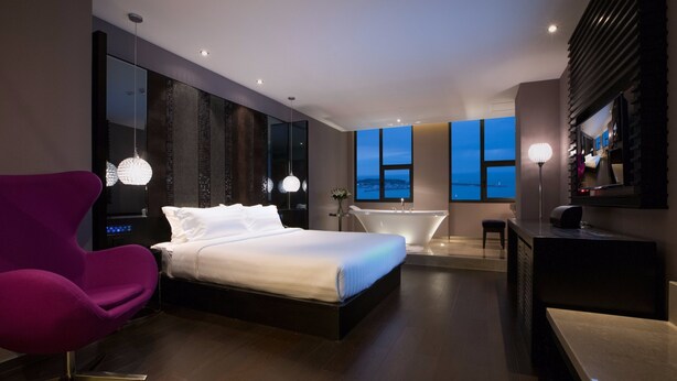 A modern hotel room with a large bed, a vanity with a mirror, a bathtub, and a view of the cityscape.