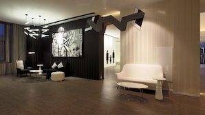 A modern living room with a white sofa, black and white patterned ottoman, and a round coffee table. A large abstract painting adorns the wall, and a unique zigzag-shaped light fixture hangs from the ceiling.