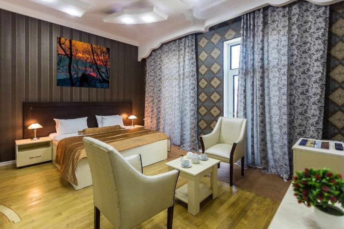 A hotel room with a large bed, a bedside table with a lamp, a desk with a chair, and a window with patterned curtains.