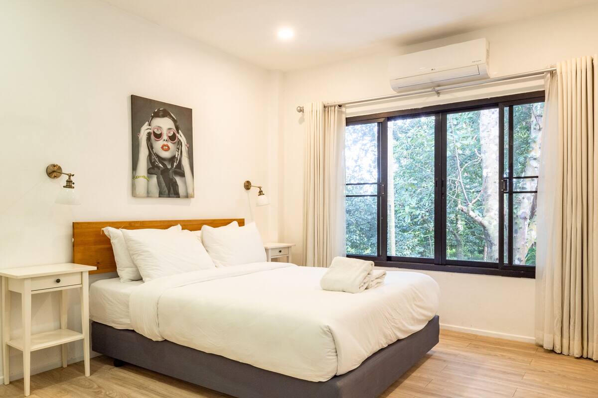 Ein Schlafzimmer mit Bett, Nachttischen, einem Gemälde, einem Fenster mit Vorhängen und einer Klimaanlage.