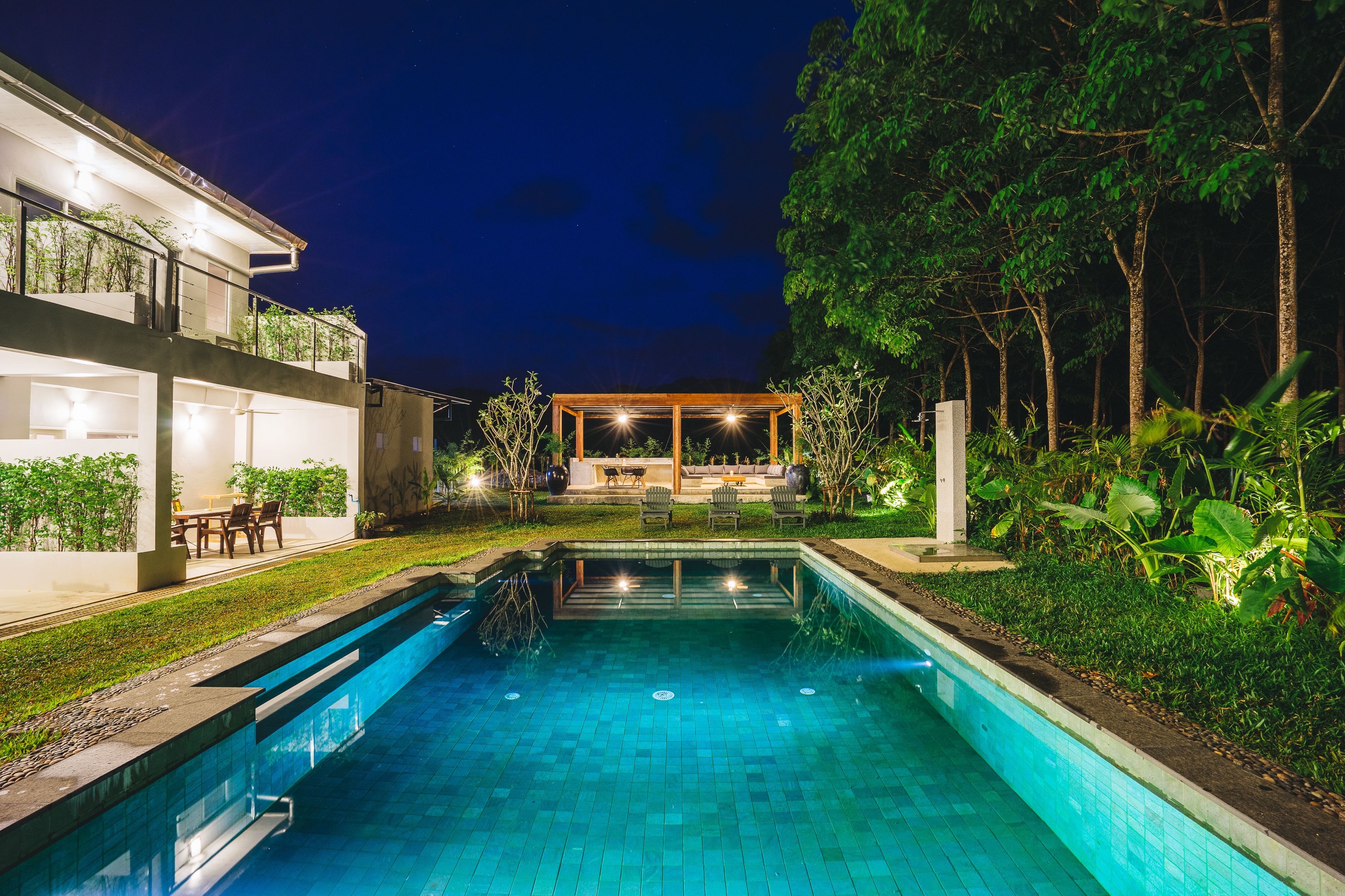 Una piscina con cubierta de madera y zona de estar, rodeada de exuberante vegetación y una casa moderna de varios niveles con iluminación exterior.