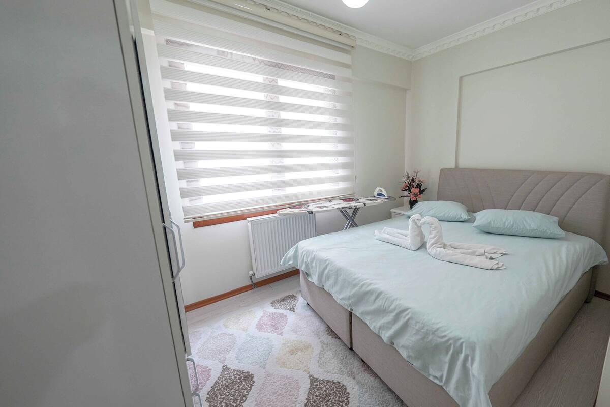 A bedroom with a bed, a radiator, a window with blinds, a small table with an ironing board, and a vase with flowers.