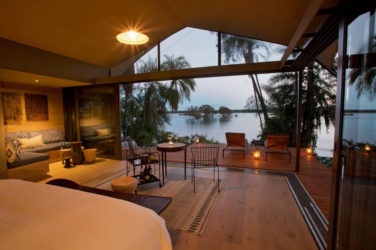 A modern bedroom with a large bed, a sofa, and a dining area with a table and chairs. There is a view of a body of water and palm trees outside the windows.