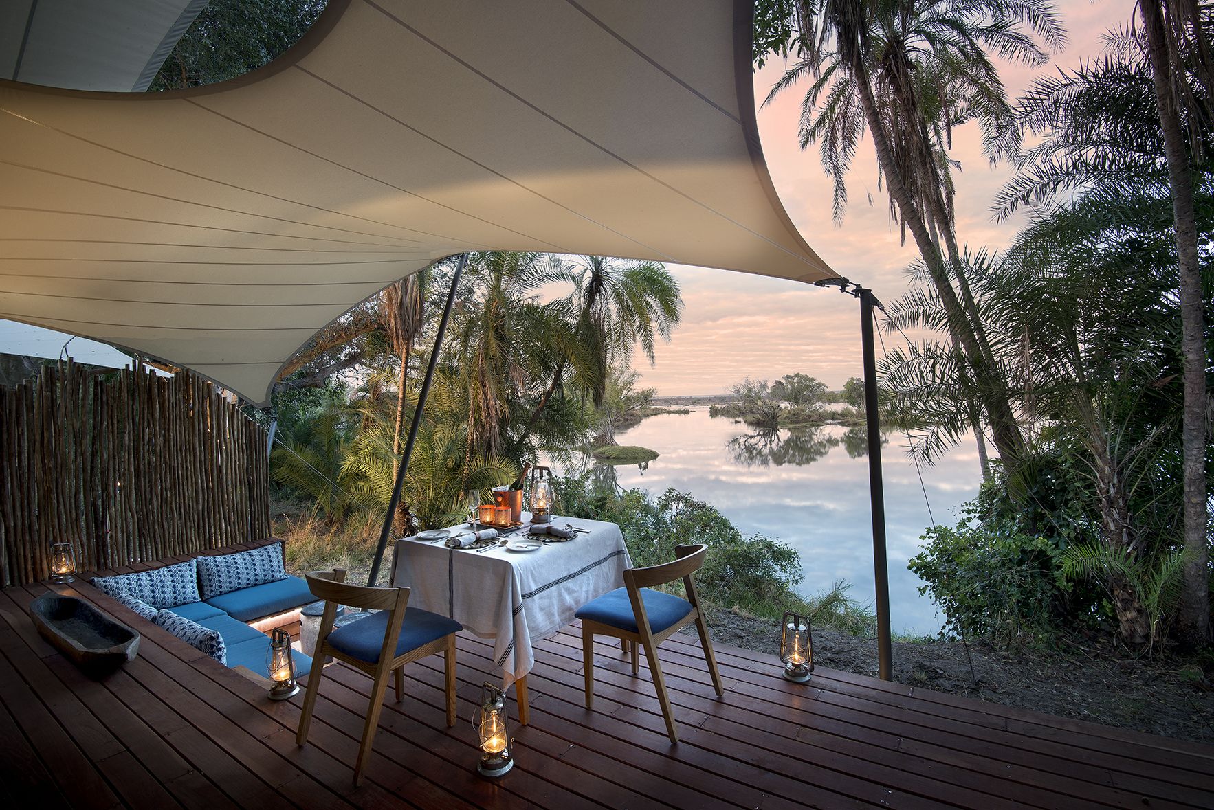 An outdoor dining area with a table set for two, surrounded by wooden deck flooring, under a large canopy.