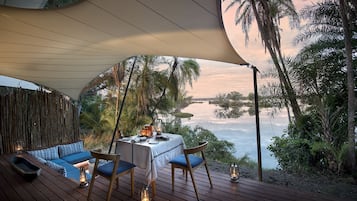 An outdoor dining area with a table set for two, surrounded by wooden deck flooring, under a large canopy.